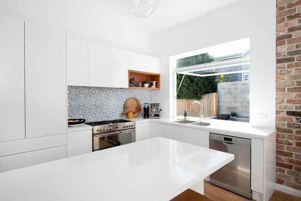 Bright contemporary white kitchen with a large central island, white flat-panel cabinetry, patterned grey splashback tiles, stainless-steel range cooker and dishwasher, and a white benchtop. A large opening window above the sink overlooks a small courtyard, while exposed red brick forms a feature wall at right.