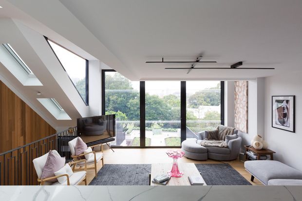 Bright contemporary living room with a large sectional sofa, lounge chairs, ottoman and coffee table, opening to a balcony or terrace through full-height glazing. The room features a dramatic sloped ceiling with multiple skylights, warm vertical timber wall panelling, dark-framed glazing, a linear ceiling fan, and a partial view of a kitchen island or benchtop in the foreground.