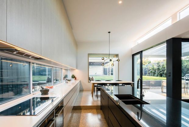Bright contemporary kitchen with a long wall of integrated cabinetry and display glazing, a central island with dark cabinetry and a black sink, and a dining area beyond. Full-height and clerestory glazing bring in abundant daylight, with views toward a landscaped outdoor area. Polished light terrazzo-style flooring and pale cabinetry create an airy, refined interior.