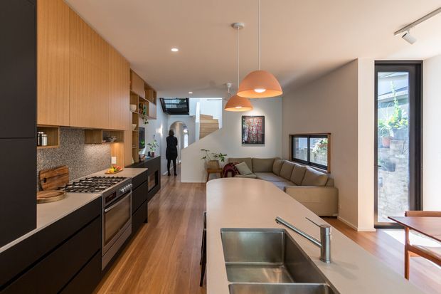 Open-plan contemporary kitchen and living area with a long light stone island, stainless-steel sink, black lower cabinetry, warm timber overhead cabinetry and flooring, and a textured stone backsplash. The kitchen opens to a compact lounge with a sectional sofa, while pendant lights, track lighting, and full-height glazing brighten the space.