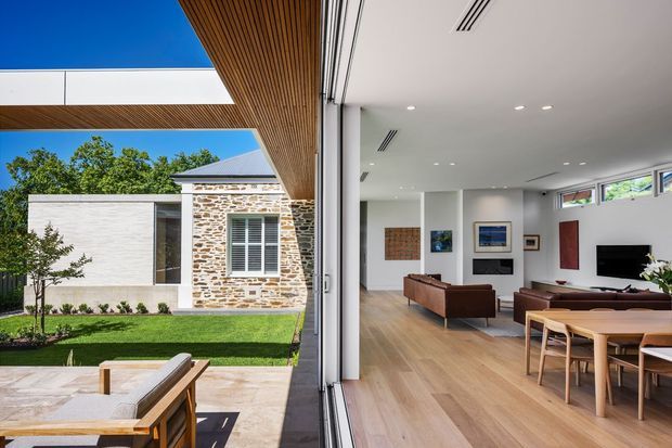 A bright contemporary open-plan living and dining space connected to a landscaped courtyard through expansive sliding glass doors. The interior features pale timber flooring, white walls, high-level clerestory windows, streamlined linear air vents, brown leather seating, a dining table, and wall-mounted television. Outside, a stone-paved terrace, lawn, timber-lined soffit, skylight opening, and a facade combining white masonry with exposed stonework are visible.