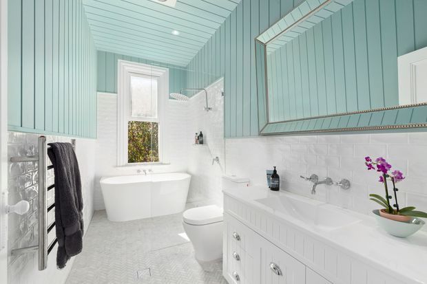 Bright, refined bathroom with a freestanding white bathtub beneath a window, a white toilet, double vanity with integrated cabinetry, wall-mounted chrome taps, white subway-tile walls, and pale aqua vertical wall panelling. A large framed mirror, heated towel rail, recessed shelving, and subtle patterned floor tiles complete the fresh coastal-inspired scheme.