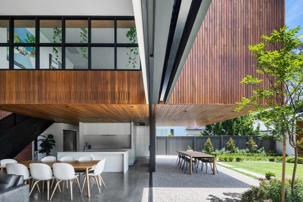 A contemporary indoor-outdoor living and dining area beneath a dramatic cantilevered upper level. The open kitchen and dining space features a long timber-lined soffit, full-height black-framed glazing, white cabinetry, a light dining table with white chairs, and polished grey flooring. Beyond the glazing is a gravel courtyard with an outdoor dining table, lawn, boundary fencing, and mature planting. Vertical timber cladding and strong black structural framing define the modern architecture.