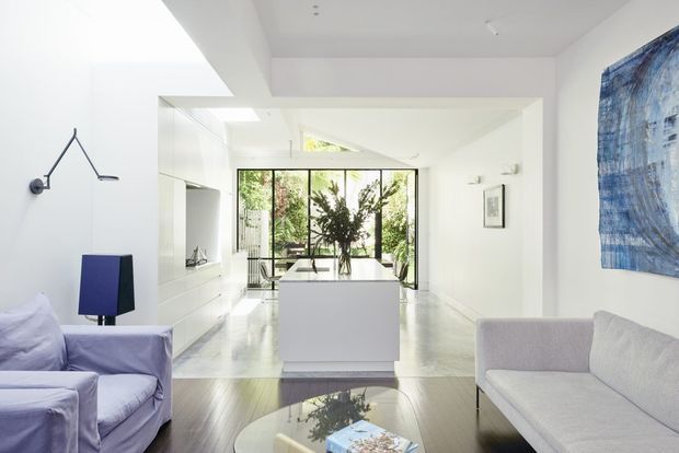 Bright contemporary open-plan interior combining a living area and kitchen. A white kitchen island anchors the space, with extensive floor-to-ceiling glazing opening toward greenery, polished light flooring, white walls and ceiling, and minimalist pale upholstered seating. A large blue artwork and blue-purple accent lighting add colour.