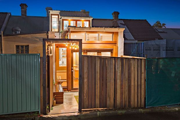 Twilight exterior view of a narrow urban residential extension behind a tall timber and corrugated-metal boundary fence. The renovated multi-level facade features warm timber-framed glazing, a projecting upper storey, roof terraces or balconies, and illuminated interiors visible through the open pedestrian gate and windows.