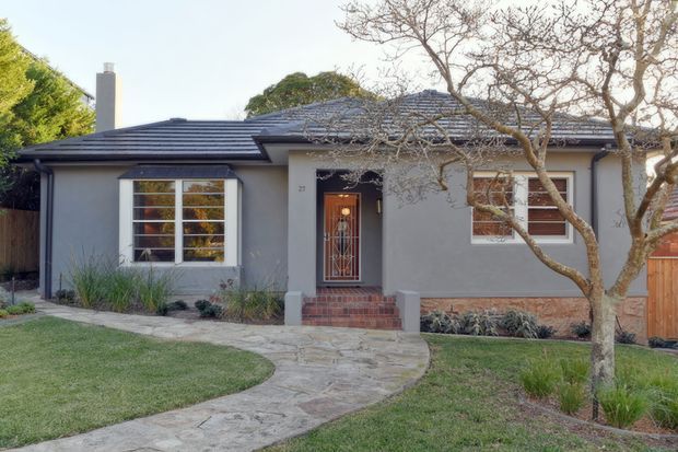 Front exterior view of a single-storey suburban house with a low-pitched dark tiled roof, pale rendered walls, brick accents around the base and entry steps, white-framed windows, and a recessed front door. A curved concrete pathway leads through a landscaped lawn to the entrance.