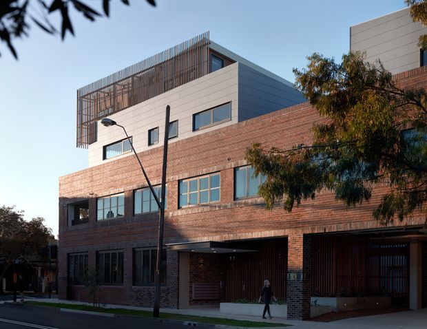 Street-facing view of a contemporary multi-storey brick building in an urban setting. The facade combines extensive red-brown brickwork with pale horizontal cladding, dark-framed windows, recessed ground-floor entries, and a rooftop terrace screened by vertical slats. Street trees and paving are visible in the foreground.