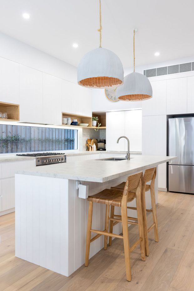 Bright contemporary kitchen featuring a large white island with an integrated sink and seating for four, white flat-panel cabinetry, pale timber flooring, open timber display niches, stainless-steel appliances, and two oversized woven pendant lights.