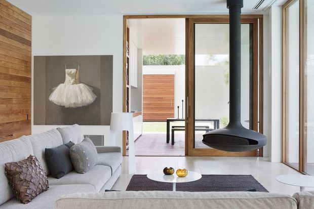 Bright contemporary living room with a pale upholstered sectional sofa, layered cushions, a low coffee table, and a suspended black wood-burning fireplace. Full-height glazing and timber-framed sliding doors open toward a timber-clad courtyard or exterior wall. A timber feature wall and large framed artwork add warmth to the otherwise light, minimal interior.