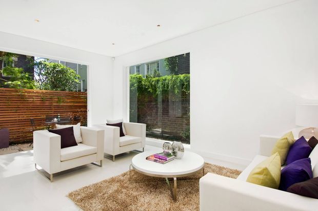Bright, minimalist living room with white walls, a white ceiling, large windows, and a white upholstered sofa and armchairs arranged around a round coffee table. A warm brown shag rug and purple and yellow cushions add contrast, while greenery and a horizontal timber privacy screen are visible outside.