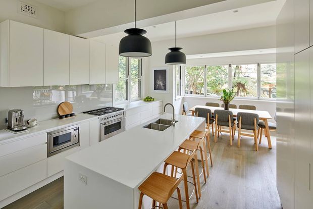 Bright open-plan white kitchen with extensive handleless cabinetry, a large white island with integrated sink and seating, stainless-steel appliances, and two black pendant lights. The kitchen opens directly into a dining area with a timber dining table and upholstered chairs, with abundant windows overlooking greenery. Light timber flooring and a restrained monochromatic palette create an airy, clean, contemporary space.