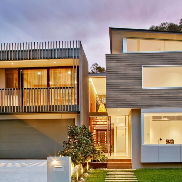 Contemporary multi-storey residential facade photographed at dusk, featuring two interlocking flat-roofed volumes with dark horizontal cladding, extensive warm-lit glazing, a slatted balcony screen, open stairwell, garage, and a landscaped front path.