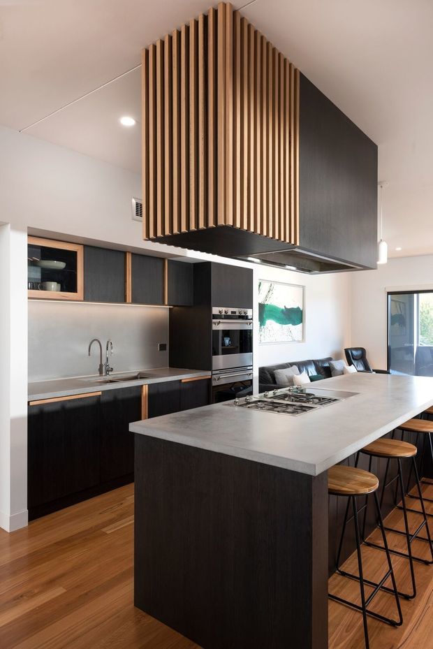 A contemporary open-plan kitchen with a large island and integrated gas cooktop, dark timber cabinetry, stainless-steel appliances, and a pale stone benchtop. A dramatic suspended rangehood is clad in vertical timber battens, creating a strong architectural feature. Timber flooring, recessed lighting, pendant lights, bar stools, and an adjacent living area are visible.