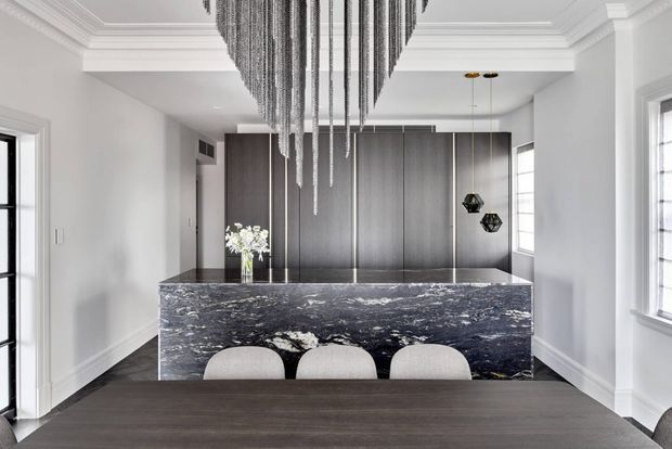 A refined contemporary kitchen and dining area viewed from the dining table. The space features a large dark natural-stone island with waterfall sides, full-height dark timber cabinetry, white walls and elaborate decorative ceiling mouldings. A dramatic cascading linear pendant hangs above the island, while three light upholstered stools sit at the island. Black-framed glazing and pendant lights add contrast.