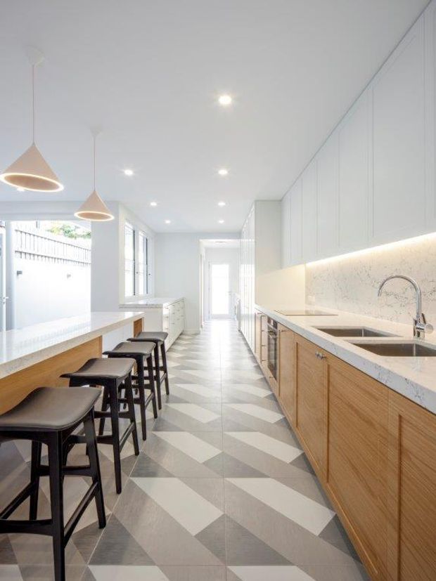 Bright, narrow contemporary kitchen with a long timber island and black stools on the left, full-height pale cabinetry and a stone benchtop with integrated sink on the right. Geometric grey-and-white floor tiles, subtle under-cabinet lighting, pendant lights, and abundant glazing create a clean, streamlined space.