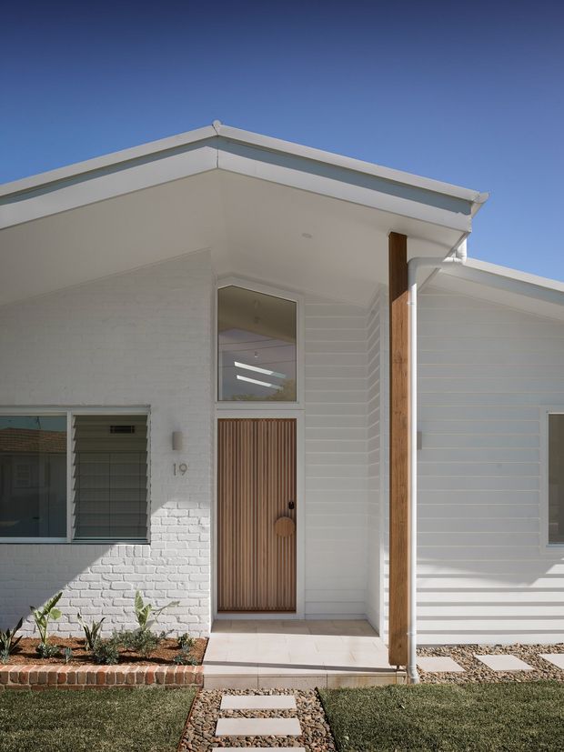 Front exterior view of a single-storey residence with a white painted brick and horizontal weatherboard facade, broad shallow-pitched roofline, timber-lined entry door, timber post, louvred window, and a simple landscaped front path with stepping stones.