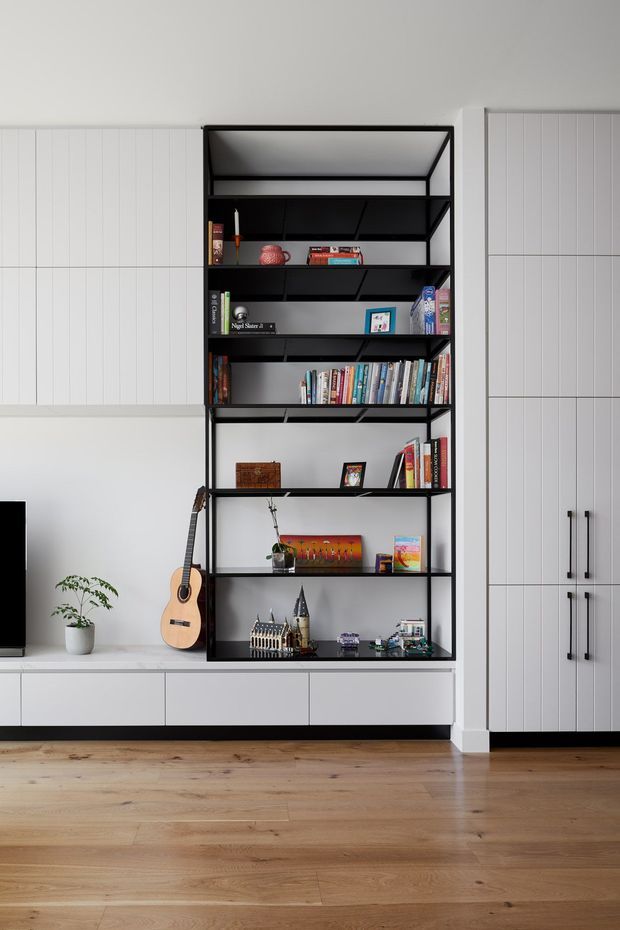 A bright, minimalist living room wall featuring a floor-to-ceiling black metal open shelving unit filled with books and decorative objects, integrated between white built-in cabinetry. White vertical panelled cabinetry with black handles surrounds the shelving, with low white drawers below. A timber acoustic guitar leans beside the unit, and pale timber flooring extends across the room. A partial television and potted plant are visible at the left edge.