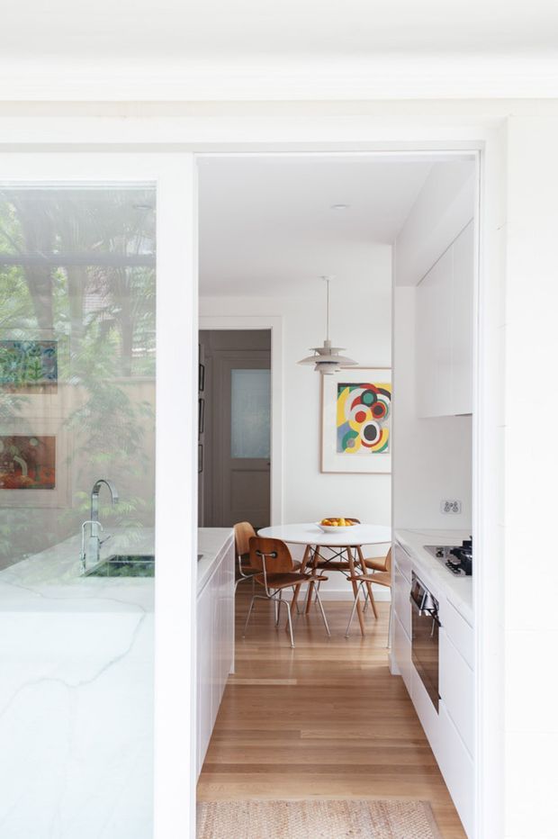 Bright, narrow contemporary kitchen viewed through a wide opening toward a compact dining area. The kitchen has white full-height cabinetry, white benchtops, integrated appliances and a gas cooktop, with warm timber flooring continuing into the dining space. A round white dining table, mid-century style timber chairs, pendant lighting and colourful abstract artwork add visual interest. Glazing at left reflects or reveals greenery and a pale stone surface.