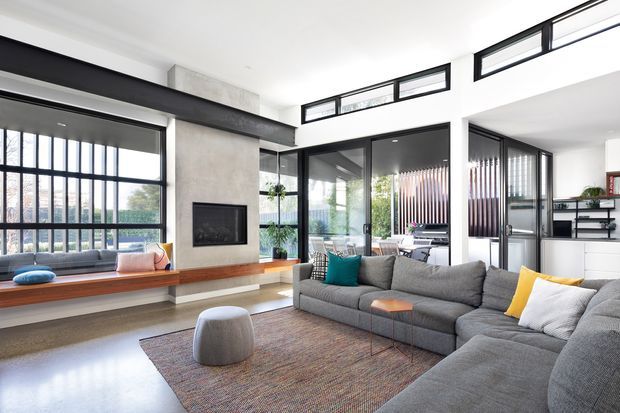 Bright contemporary living room with a large grey sectional sofa, textured area rug, and low upholstered ottoman. A long timber window seat runs beneath expansive black-framed glazing, while a concrete-look fireplace surround with built-in firebox forms a feature wall. Black-framed glass doors and internal partitions open toward a secondary workspace or dining area, with vertical screening and shelving visible beyond.
