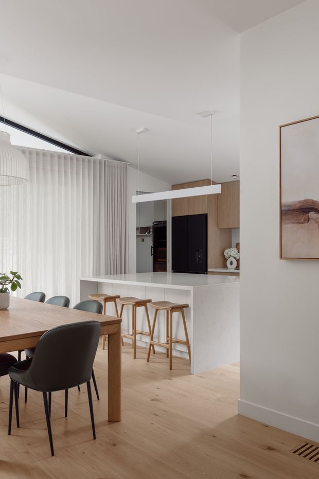 Bright, refined open-plan dining and kitchen interior. A light timber dining table with dark upholstered chairs occupies the foreground, while a white stone island with timber stools separates the dining area from the kitchen. The kitchen features light timber cabinetry, a dark integrated appliance wall, white surfaces, sheer floor-to-ceiling curtains, pale timber flooring, and minimalist linear pendant lighting. Neutral walls and restrained artwork create a calm contemporary atmosphere.
