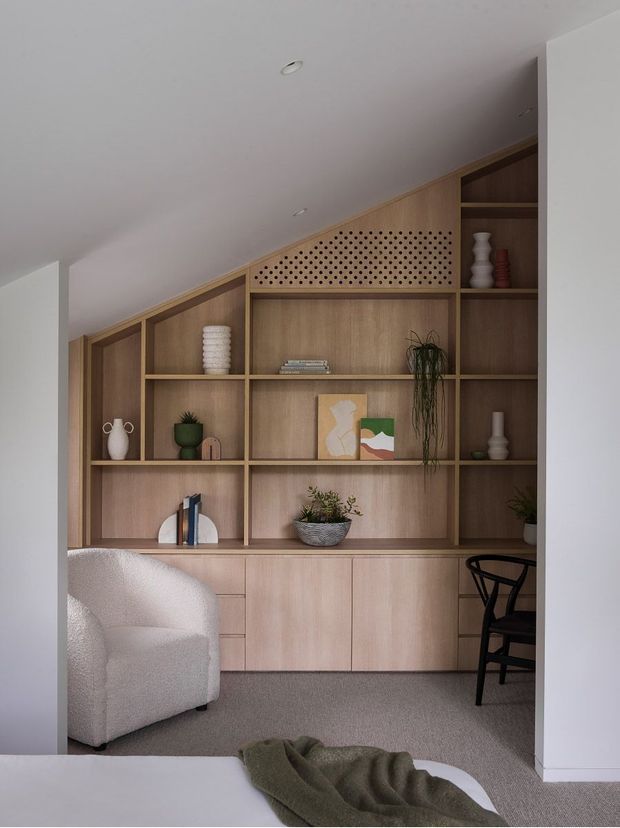 A calm, softly lit interior nook featuring a full-height custom timber built-in shelving and cabinetry wall beneath a sloping ceiling. The joinery combines open display shelves, closed lower cupboards, and a decorative perforated timber panel. Neutral upholstered seating, a dark timber chair, muted ceramics, plants, and minimalist artwork create a warm contemporary reading or living area.