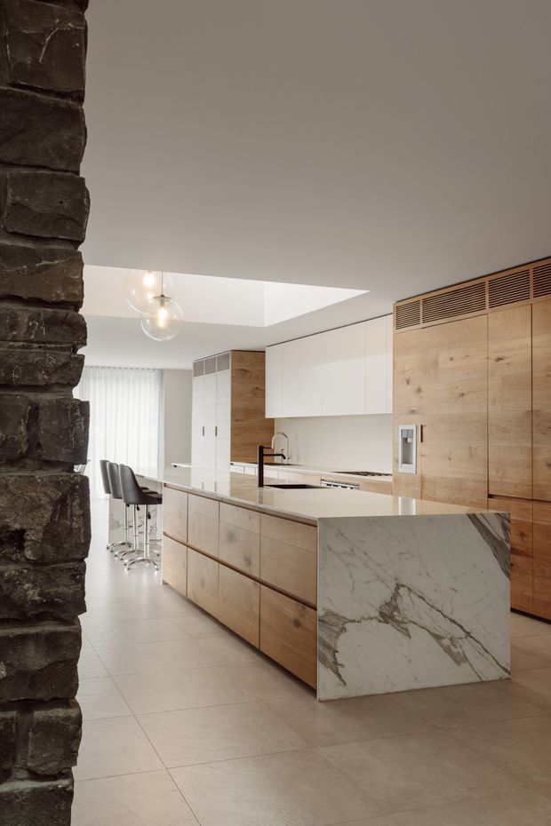 Contemporary kitchen viewed through a dark exposed stone or brick opening. The room features a large timber island with a prominent veined white stone waterfall end, pale tiled flooring, white overhead cabinetry, full-height timber joinery with integrated ventilation grilles, a black tap, bar stools, and pendant globe lighting. A broad ceiling opening or skylight brings light into the kitchen.