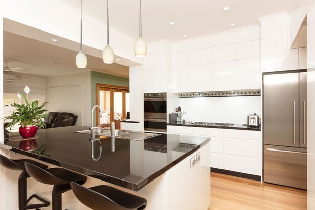 A bright, spacious contemporary kitchen with a large dark stone island and integrated sink, white full-height cabinetry, stainless-steel appliances, timber flooring, and three pendant lights. Bar stools line the island, while an open living area and dining or entry space are visible beyond.