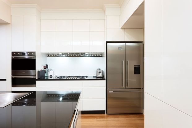 Bright contemporary kitchen with extensive white flat-panel cabinetry, integrated stainless-steel appliances, a black reflective island benchtop, a gas cooktop and rangehood, and warm timber flooring. The room has a clean, streamlined appearance with subtle decorative detailing above the cooktop.