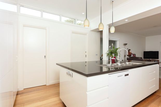 Bright contemporary white kitchen with a large central island, integrated sink and dishwasher, dark stone benchtop, white shaker-style cabinetry, timber flooring, pendant lights, and high clerestory windows. An adjacent living area is visible beyond the kitchen.