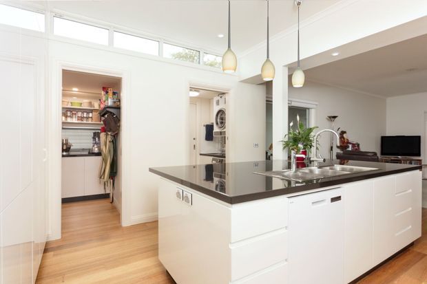 Bright open-plan contemporary kitchen with a large white island, dark stone benchtop, integrated sink and dishwasher, pendant lighting, and timber flooring. A walk-in pantry is visible to the left, with a laundry area and living space beyond the kitchen. High clerestory windows and white cabinetry create an airy, clean interior.