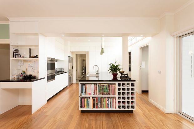Bright, open-plan contemporary kitchen and living area with white built-in cabinetry, black benchtops, warm timber flooring, and a central island featuring open bookshelves and wine storage. The space includes a sink, pendant light, integrated appliances, and large glazed doors/windows, with a hallway and adjacent rooms visible beyond.