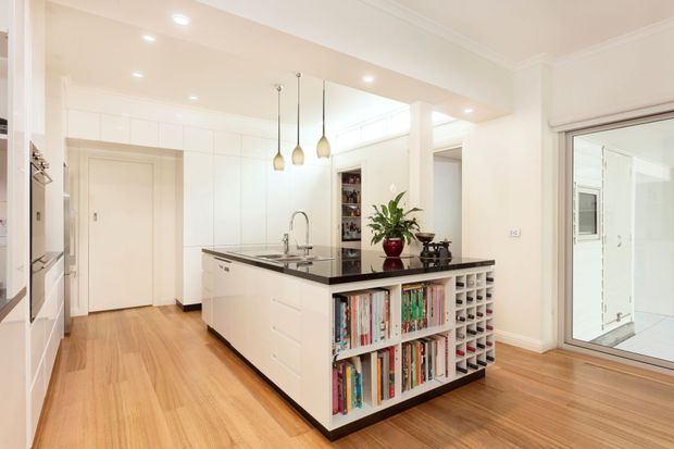 Bright, spacious contemporary kitchen centred around a large white island with a dark stone benchtop, integrated sink, open cookbook shelving and wine storage. White full-height cabinetry, timber flooring, recessed ceiling lights and three pendant lights create a clean, warm, functional interior. A glazed opening connects to an adjacent bright room.