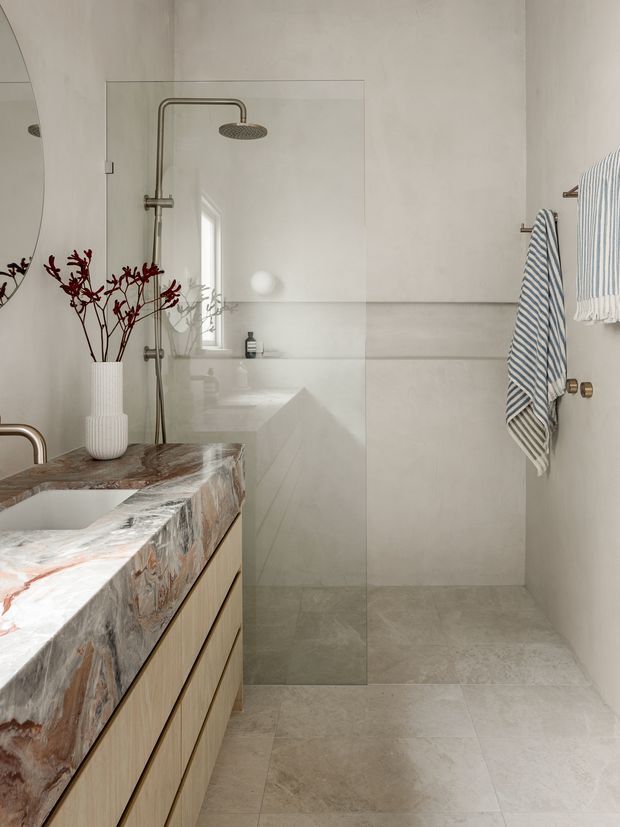 Contemporary bathroom with a walk-in glass shower, brushed metal shower fittings, pale large-format wall and floor tiles, and a floating timber vanity with a dramatic natural stone benchtop. A sculptural white vase with branches and striped towels add restrained decorative detail.