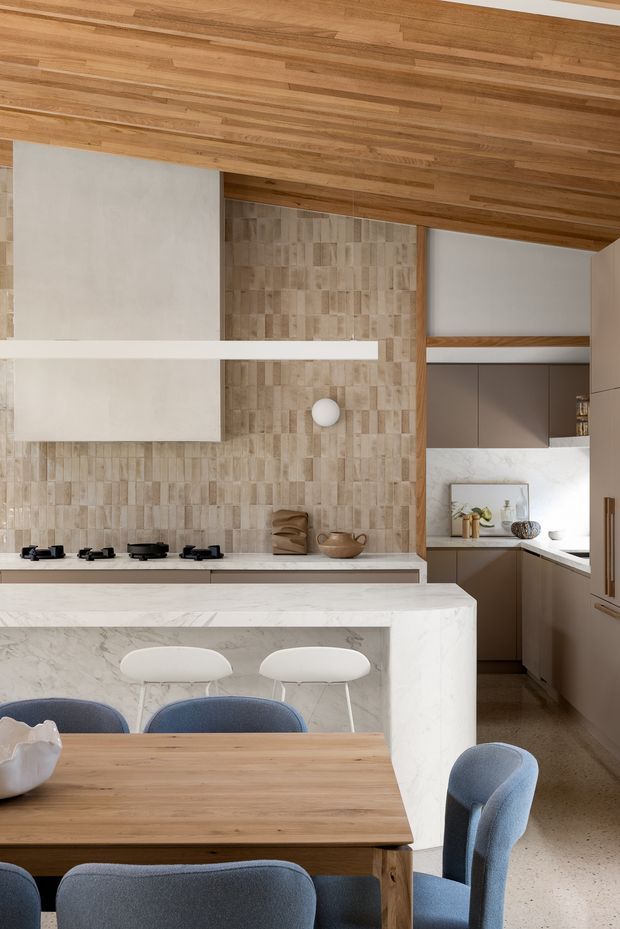A warm, contemporary open-plan kitchen and dining area. The kitchen features a large white marble or engineered-stone island with integrated seating, taupe-brown cabinetry, a textured beige tile backsplash, white cabinetry, and a timber-lined ceiling. A timber dining table with blue upholstered chairs occupies the foreground, while a secondary kitchen workspace is visible to the right.