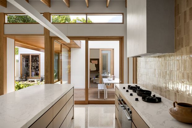 Bright contemporary kitchen with a large light stone island, pale cabinetry, matching stone benchtops, a long tiled splashback, and timber-framed glazing and doors. Exposed timber roof beams and clerestory windows bring warmth and abundant natural light. The kitchen opens toward a dining area and a planted courtyard beyond.