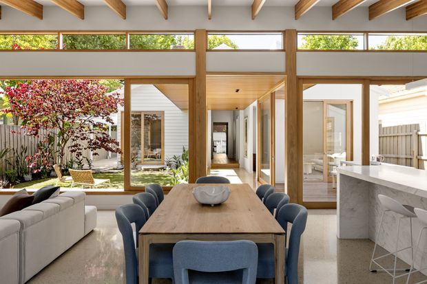 A bright open-plan dining and kitchen interior with a large timber dining table, upholstered blue dining chairs, and a pale sectional sofa. Full-height timber-framed glazing and internal doors open toward a landscaped courtyard, while exposed timber beams span the ceiling. The kitchen features a pale stone island with integrated seating. The space has a warm, refined contemporary character with strong indoor-outdoor connection.