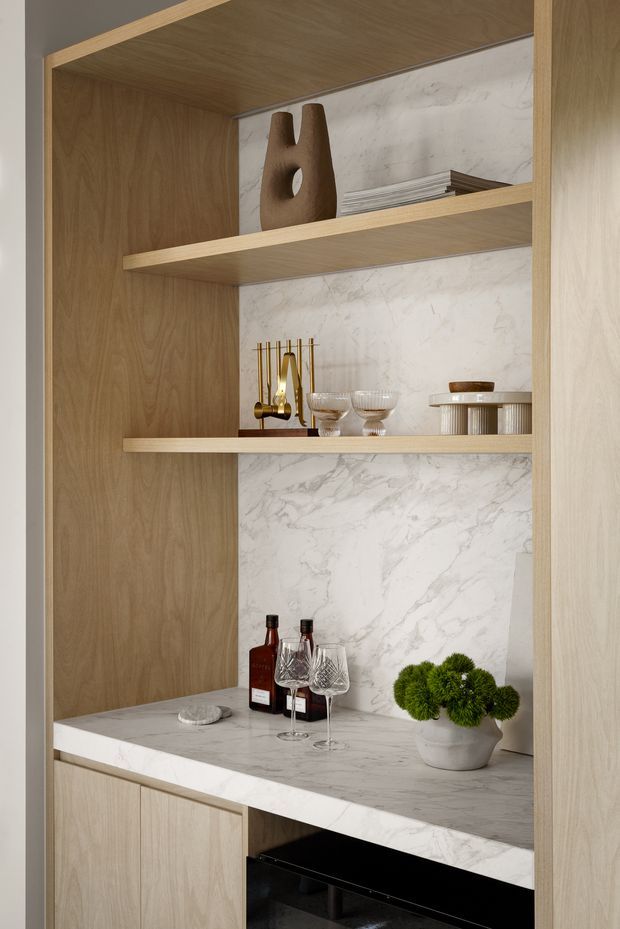 A close-up of a refined built-in bar or drinks cabinet with light timber shelving and cabinetry, a white veined stone backsplash and benchtop, and curated glassware, bottles, decorative ceramics, and brass accessories. The lower section includes dark appliance or storage space. The composition is warm, minimalist, and carefully styled.