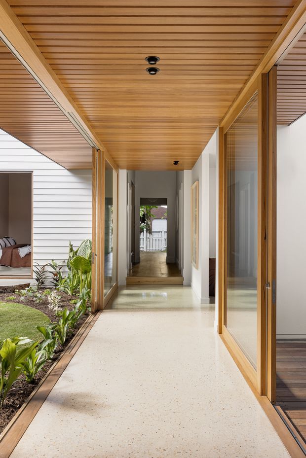 A bright residential hallway or breezeway connecting interior rooms around a planted courtyard. The passage has a warm timber-lined slatted ceiling, extensive timber-framed glazing and doors, pale walls, and a light terrazzo-like floor. A garden strip and adjoining living space are visible to the left, with a corridor extending toward an exterior gate in the distance.