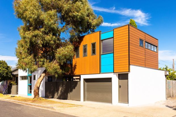 Street-facing view of a compact contemporary multi-storey residence with a white rendered lower level, bold orange timber-look horizontal cladding on the upper storey, and bright turquoise glazing or panels between projecting volumes. The house includes a single garage, narrow entry door, elevated upper-level form, and a mature street tree in front. Strong geometric composition and vivid contrasting colours define the facade.