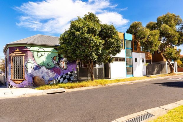 Street-level exterior view of a mixed residential streetscape. A low older building with a corrugated metal roof and vivid mural artwork sits beside a narrow contemporary multi-storey residence with white walls, dark vertical cladding, and prominent blue-tinted glazing. Mature street trees, a paved footpath, curb, roadway, and stormwater drain are visible under bright sunlight.