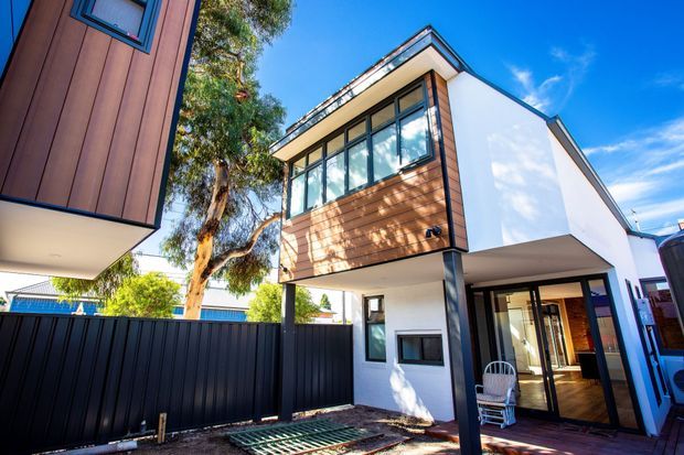 Exterior view of a contemporary multi-storey residence with a projecting upper-level volume, white rendered walls, warm timber-look vertical cladding, extensive glazed windows, and a covered entry or outdoor living area. The property sits in a compact urban or suburban setting with dark boundary fencing and paved surfaces.
