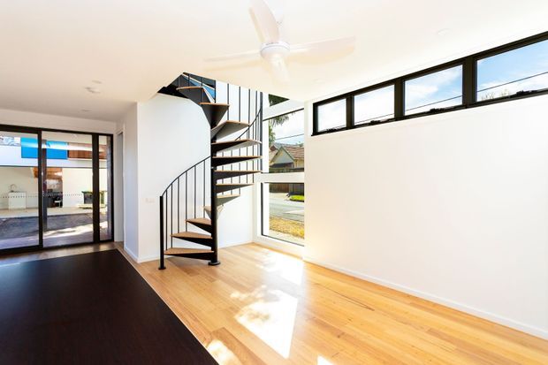 Bright contemporary interior featuring a compact black metal spiral staircase with timber treads, light timber flooring, white walls, clerestory-style horizontal windows, and extensive glazing opening toward an outdoor area. A dark timber-toned floor section contrasts with the pale timber floor, while a ceiling fan and minimal finishes create an airy, clean space.
