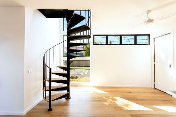 Bright, minimalist interior room featuring a sculptural black metal spiral staircase, pale timber flooring, white walls, high horizontal windows, and an open door. Sunlight creates warm highlights across the floor, while an outdoor garden and paving are visible through the glazing. A ceiling fan is mounted overhead.