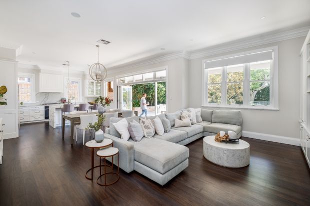 Bright open-plan living room with a large light-grey sectional sofa, round textured coffee table, dark timber flooring, white walls and detailed ceiling cornices. A contemporary white kitchen with island seating and pendant lighting is visible at the rear, along with expansive windows and glazed doors opening to greenery.