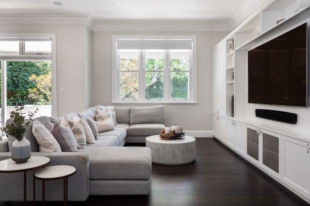 Bright, refined living room with a large light-grey sectional sofa, layered neutral cushions, a round stone coffee table, and dark timber flooring. White built-in entertainment cabinetry spans the right wall, incorporating open shelving, a wall-mounted television, soundbar, and concealed storage. The room features pale walls, decorative ceiling cornices, plantation-style window trim, and abundant garden outlook through the windows.