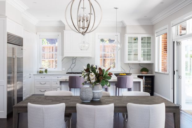 Bright, refined kitchen-dining space with white shaker-style cabinetry, a dark stone or stone-look island and dining table, stainless-steel appliances, marble-look splashback, and upholstered dining chairs. A large spherical metal chandelier is suspended above the table, while abundant windows and glazed doors provide natural light. The room has classic detailing including decorative cornices and paneled cabinetry, with a contemporary, polished fit-out.