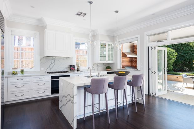 Bright, refined white kitchen with detailed panelled cabinetry, marble-look surfaces, a central island with four upholstered stools, dark timber flooring, and large glazed doors opening to an outdoor patio. A brick-lined built-in barbecue or fireplace feature is visible near the opening.