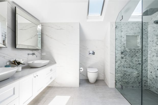 Bright contemporary bathroom with a double vanity, white panelled cabinetry, pale grey benchtop, large white marble-look wall slabs, grey floor tiles, a wall-hung toilet, and a frameless glass shower enclosure lined with patterned grey tiles. A skylight provides strong natural light, and a mirrored cabinet reflects the room.