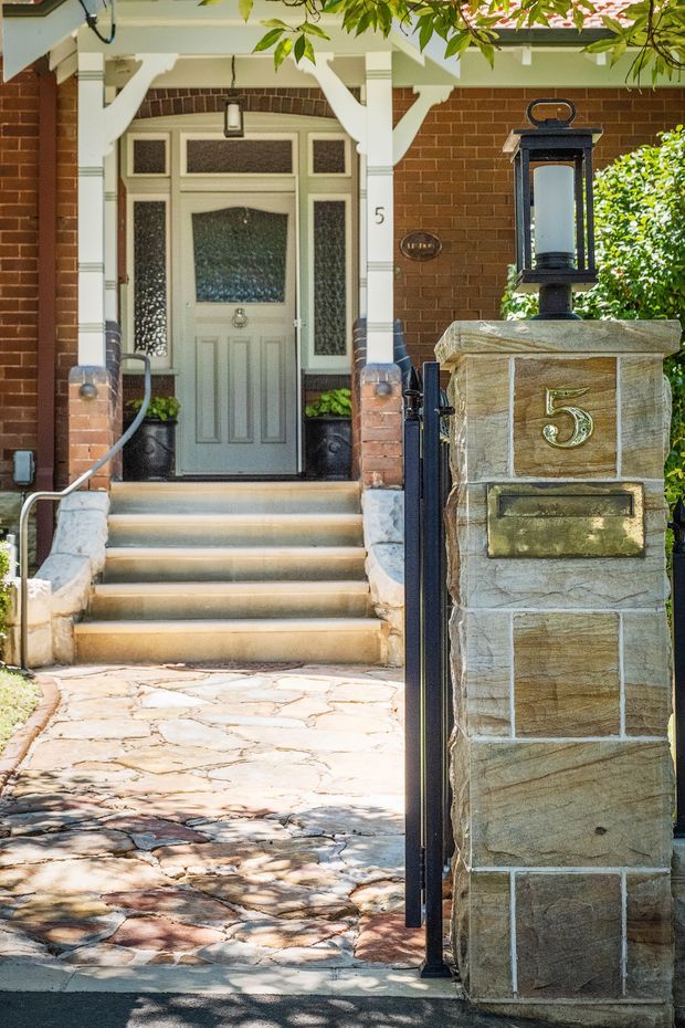 A close exterior view of a traditional residential front entry. A pale green glazed front door with sidelights and transom is framed by white decorative timber trim against a red brick facade. A short flight of steps leads up from a paved walkway, with a sandstone-clad pillar featuring a large lantern, house number 5, and mailbox in the foreground. Garden foliage partially frames the entry.