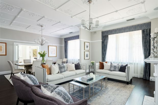 A bright, refined formal living and dining room with ornate decorative ceiling panels, high ceilings, large curtained windows, pale upholstered sofas, patterned accent cushions, dark timber flooring, and a glass coffee table with a polished metal frame. A dining table and chairs are visible in the adjoining area, while a classical fireplace mantel appears at right.